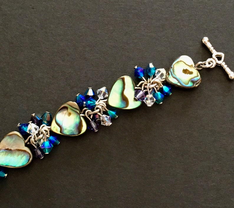 May include: A bracelet featuring three heart-shaped abalone shell charms interspersed with clusters of faceted blue, purple, and clear crystal beads. The jewelry piece is finished with a silver-toned metal toggle clasp shaped like a small heart.