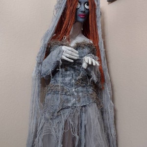 May include: A handmade, spooky bride doll with a blue face, red hair, and a tattered gray dress. The doll has a veil and is decorated with moss and other natural elements. The doll is a unique piece of art.