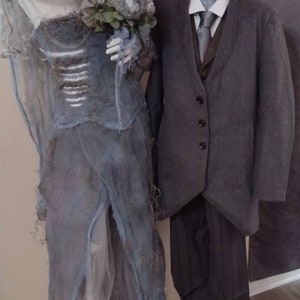 May include: A Halloween-themed display featuring two mannequins. One is dressed as a ghostly bride in a tattered blue gown, holding a bouquet. The other mannequin is dressed in a gray suit with a white shirt and tie. A large clock is in the background.