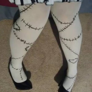 May include: White tights with black stitched detailing, resembling a doll's design. The tights feature a heart shape near the ankle and are paired with black high-heeled shoes. A black and white striped skirt is visible at the top.