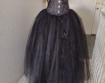 May include: A dark gray corset with a floral pattern and a black tulle skirt. The corset has a lace-up front and decorative trim. A black spider embellishment is attached to the skirt. The outfit is displayed on a mannequin.