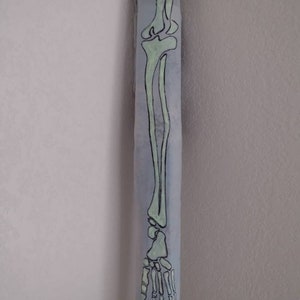 May include: A light blue, rectangular decorative item with a hand-drawn illustration of a human leg skeleton. The bones are depicted in a light green color with black outlines. The item is positioned in a corner.