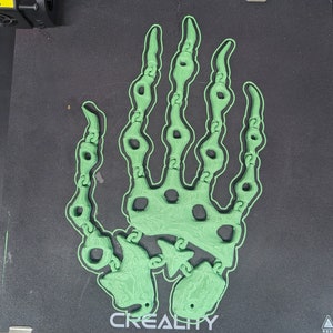 Op de afbeelding: Een 3D-geprint, groen skelet handontwerp op een zwart oppervlak. De hand is gedetailleerd met botachtige structuren en is waarschijnlijk een decoratief of artistiek stuk. De basis heeft het woord "CREALITY" erop gedrukt.