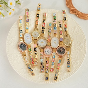 Może przedstawiać: Kolekcja złotych zegarków na rękę z ozdobnymi bransoletkami z zawieszkami. Tarcze zegarków są w różnych kolorach, w tym białym, czarnym i różowym. Bransoletki zdobią kolorowe, miniaturowe wzory. Zegarki ułożone są na białym, wzorzystym talerzu.