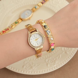 May include: A gold-toned watch with a white face and crystal accents is worn with a matching bracelet. A second gold watch with a patterned band is in the background. The watch on the wrist has the word "DOUKOU" on the face.