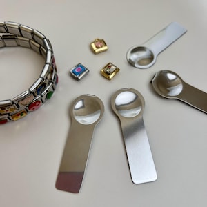 Könnte beinhalten: Eine Sammlung von Schmuckstücken, darunter ein silbernes Armband mit bunten Emaille-Anhängern, kleine dekorative Anhänger und mehrere silberne Löffelwerkzeuge. Die Anhänger sind quadratisch und goldfarben, mit verschiedenen Designs. Die Löffelwerkzeuge haben eine abgerundete Schale und einen flachen Griff.