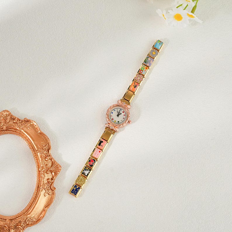 May include: A gold-coloured wristwatch with a white face and a decorative strap. The strap has colourful, square-shaped links with various designs. The watch has a rose gold-coloured bezel and a delicate, feminine design. The watch is a fashion accessory.