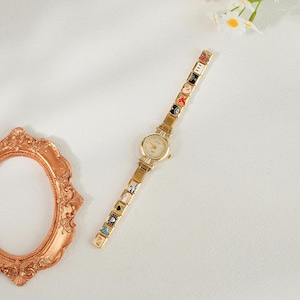 May include: Gold-coloured wristwatch with a round face and a decorative band featuring colourful enamel charms. The watch has a classic design with a gold-coloured case and a delicate, patterned band. The charms include various symbols and designs.