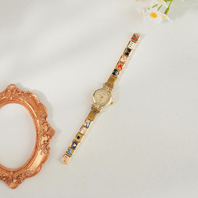 May include: Gold-coloured wristwatch with a round face and a decorative band featuring colourful enamel charms. The watch has a classic design with a gold-coloured case and a delicate, patterned band. The charms include various symbols and designs.
