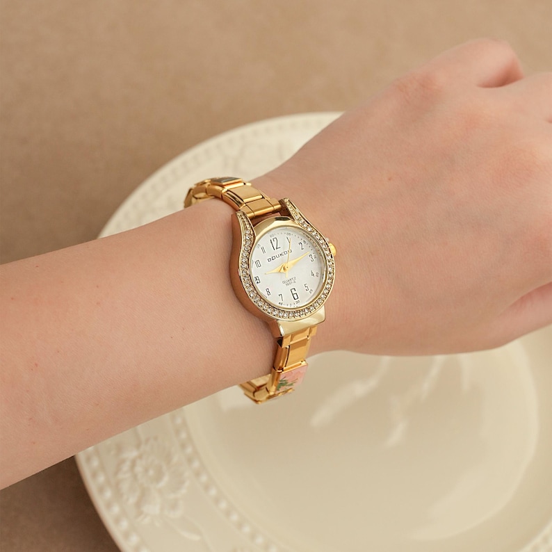 May include: Gold-toned wristwatch with a white face and a bezel encrusted with small, clear crystals. The watch has a gold band and is worn on a wrist. The watch face displays the numbers 1 through 12.