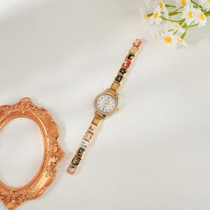 May include: A gold-coloured wristwatch with a white face and a bezel set with small, clear stones. The watch strap is made up of decorative, colourful rectangular links. The watch is on a white background with a gold-coloured picture frame and white flowers.