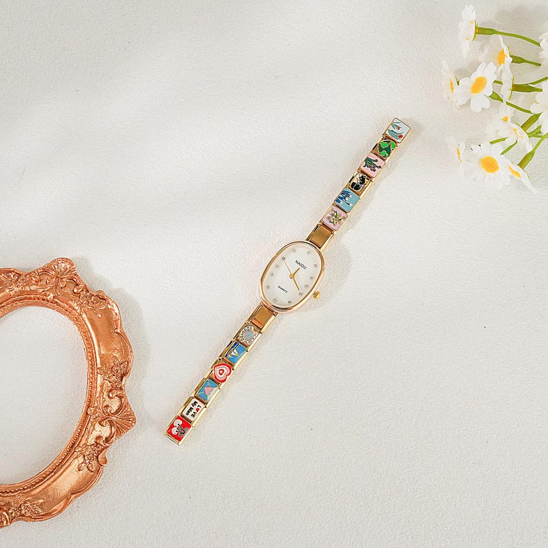 May include: Gold-coloured wristwatch with an oval white face and a patterned strap. The strap has colourful, square designs. The watch is on a white background with a decorative gold frame and white flowers.