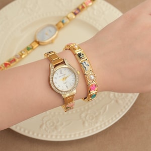May include: A gold watch with a white face and crystal detailing is paired with a matching bracelet. A second gold watch with a patterned strap is in the background. The watch face reads "DOUKOU".