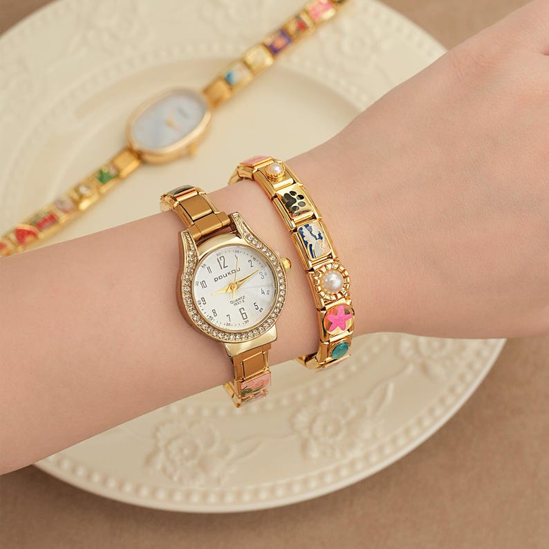 May include: A gold watch with a white face and crystal detailing is paired with a matching bracelet. A second gold watch with a patterned strap is in the background. The watch face reads "DOUKOU".