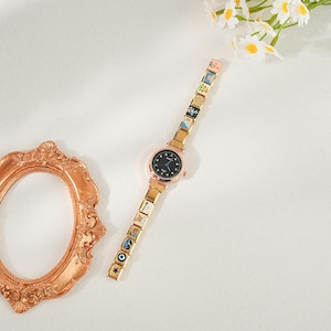 May include: A gold-coloured wristwatch with a black face and decorative charms on the strap. The watch has a round face with small crystals and a gold-coloured bezel. The strap is made of gold-coloured links, each with a small, colourful design.