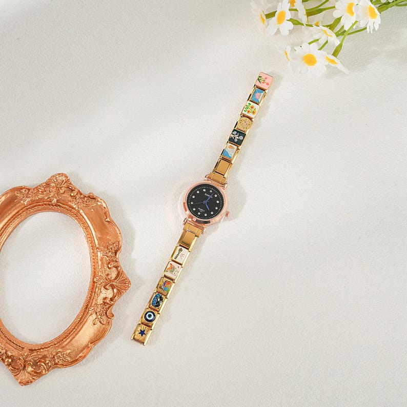 May include: A gold-coloured wristwatch with a black face and decorative charms on the strap. The watch has a round face with small crystals and a gold-coloured bezel. The strap is made of gold-coloured links, each with a small, colourful design.