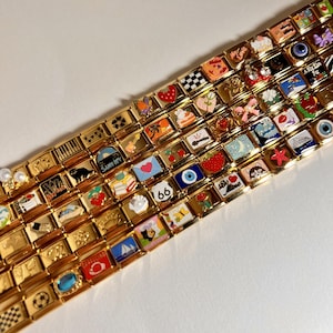 May include: A gold charm bracelet with a variety of colorful charms, including a sailboat, a strawberry, a cat, a piano, a heart, a seashell, a butterfly, a cake, a crown, a moon, a star, a red apple, a guitar, a palm tree, a suitcase, a black cat, a city skyline, a soccer ball, a checkered pattern, a red and white heart, a blue and white eye, and the number "66".