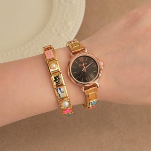 May include: A gold wristwatch with a black face and Roman numerals is shown next to a gold bracelet. The bracelet has colourful rectangular charms and pearls. The watch and bracelet are on a person's wrist.