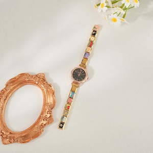 May include: A gold-coloured wristwatch with a black watch face and a strap decorated with colourful, square charms. The watch has a round face with gold hands and Roman numeral hour markers. The charms on the strap feature various designs. The watch is displayed on a white surface.