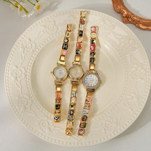 May include: Three gold-toned wristwatches with decorative charm bracelets are displayed on a white plate. The watch faces are round with Roman numerals. The bracelets feature colorful, miniature art pieces. The plate has a floral design.