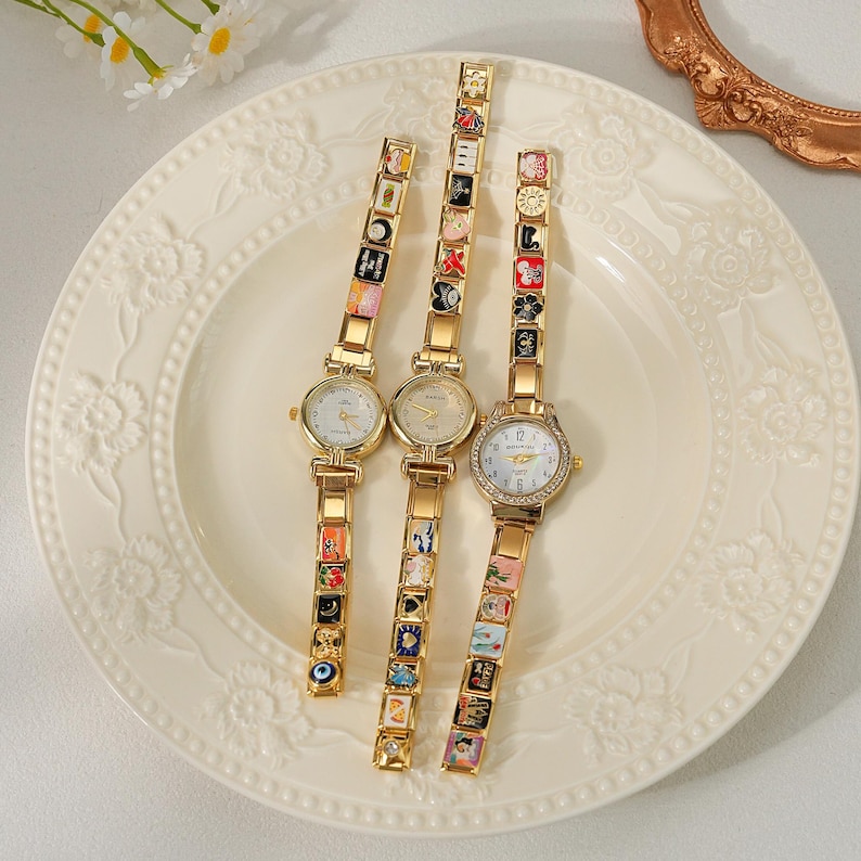 May include: Three gold-toned wristwatches with decorative charm bracelets are displayed on a white plate. The watch faces are round with Roman numerals. The bracelets feature colorful, miniature art pieces. The plate has a floral design.