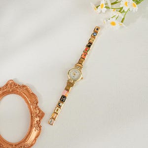 May include: A gold wristwatch with a white face and a decorative strap. The strap has colourful, square charms with various designs. The watch is set against a white background with a decorative frame and flowers.