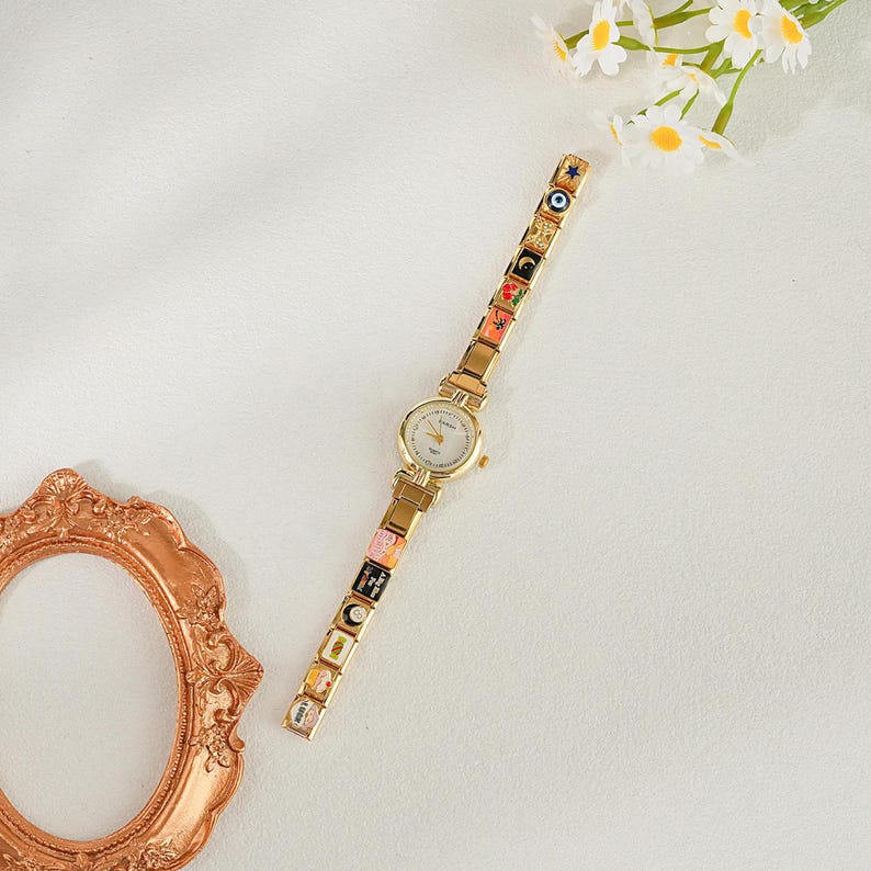 May include: A gold wristwatch with a white face and a decorative strap. The strap has colourful, square charms with various designs. The watch is set against a white background with a decorative frame and flowers.