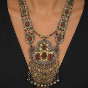 May include: A long necklace featuring oxidized silver and gold-toned metal rectangular links, each set with an oval red stone. The central pendant is a large, ornate, multi-layered piece with red stones, gold filigree, and dangling metal beads.