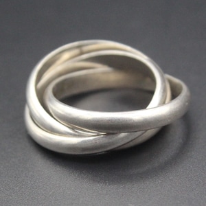 May include: A silver-colored ring with three interlocking bands. The bands are smooth and rounded, creating a seamless, intertwined design. The ring is displayed against a dark background.