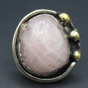 Peut inclure: Une bague en argent avec une grosse pierre ovale en quartz rose clair. La pierre est sertie dans un anneau en argent texturé avec trois petits accents dorés sur un côté. La bague est présentée sur fond gris foncé.