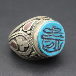 Könnte beinhalten: Ein silberner Ring mit einem blauen Stein in der Mitte. Der Stein ist mit komplizierten schwarzen Schnitzereien verziert. Der Ring ist mit silbernen Filigranen und roten und weißen Akzenten verziert.