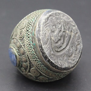 Könnte beinhalten: Antiker Silberring mit aufwendiger Drahtarbeit und einem zentralen Stein. Der Ring hat eine dunkle, gravierte Oberseite mit einem verzierten Design und einem kleinen blauen Stein. Ein einzigartiges Schmuckstück.
