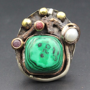 Puede incluir: Un anillo con una piedra central de malaquita cuadrada con bandas verdes, acentuada por una perla blanca, dos pequeñas piedras circulares de color rojo rosado y tres esferas doradas sobre una base de metal plateado oscuro oxidado.