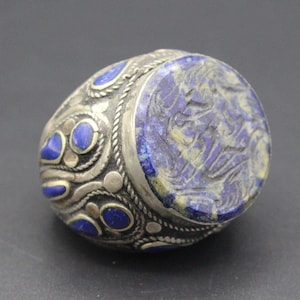 Könnte beinhalten: Ein silberner Ring mit einem blauen Lapislazuli-Stein, der in der Mitte eingesetzt ist. Der Stein ist mit komplizierten Mustern verziert und der Ring ist mit kleineren blauen Steinen verziert.