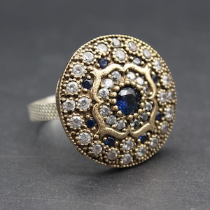 May include: A detailed, round ring with a silver band and a gold-toned, circular face. The face is adorned with numerous small, clear and dark blue gemstones, arranged in a floral pattern. The ring is set against a dark background.