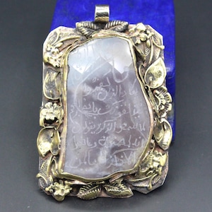 Könnte beinhalten: Ein rechteckiger Anhänger mit einem zentralen, durchscheinenden, hellvioletten Stein, der mit arabischer Kalligrafie graviert ist. Der Stein ist in einen Metallrahmen mit skulpturalen, goldfarbenen Blumen- und Blattmotiven gefasst.
