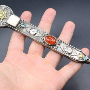 Puede incluir: Un puntero decorativo plateado y dorado con un diseño de Estrella de David, piedras preciosas rojas y azules y un dedo apuntando al final.