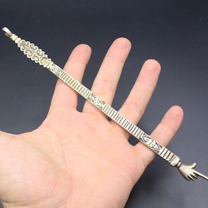 Könnte beinhalten: Ein silberfarbener Metallzeiger mit einem verzierten Design und einer nach rechts zeigenden Hand. Der Zeiger wird in einer Hand gehalten.