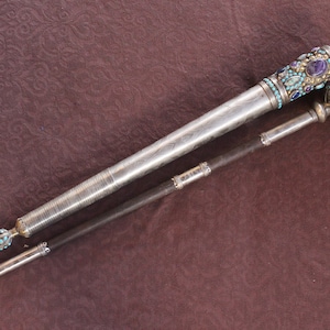 以下が含まれることがあります： 装飾的なハンドルと黒いインクカートリッジが付いた銀とターコイズのペン。ペンはキャップが端にある閉じた位置にあります。