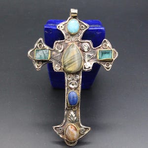 May include: A silver-toned cross pendant with inlaid gemstones. The cross features a central teardrop-shaped stone, turquoise, blue, and green rectangular stones, and an oval stone at the bottom. The cross is set against a dark background.