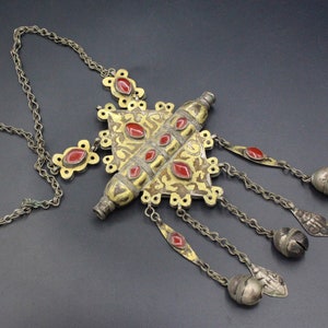 May include: Antique silver-tone necklace with a central gold-tone pendant featuring red gemstone accents. The necklace has a chain and decorative dangles with silver bells and red gemstones. The design is intricate and ornate.