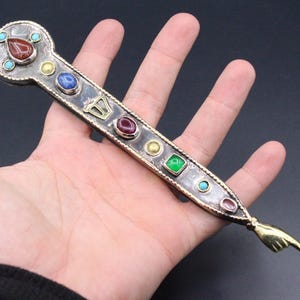 Può includere: Un pendente ornato, color argento, con una serie di pietre preziose colorate, tra cui rosso, blu e verde. Il pendente ha una forma unica con un manico decorativo e un piccolo anello nella parte superiore.