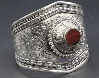 May include: A wide silver cuff bracelet with intricate engraved patterns and a central, circular, reddish-brown stone. The bracelet features detailed scrollwork and beaded edges, showcasing a vintage or ethnic design.