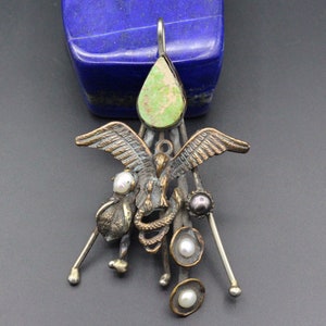 May include: A silver and bronze pendant featuring a bird with outstretched wings, a snake, and a green gemstone. The pendant is adorned with pearls and beads.