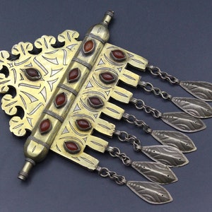 May include: A silver and gold pendant with a decorative design and red stones. The pendant has a long, cylindrical piece at the top and a series of chains with decorative leaves hanging from the bottom.