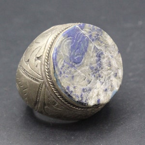 Könnte beinhalten: Ein silberner Ring mit einem ovalen Lapislazuli-Stein, der in der Mitte eingesetzt ist. Der Stein hat ein geschnitztes Design und ist von einem silbernen Band mit komplizierten Details umgeben.