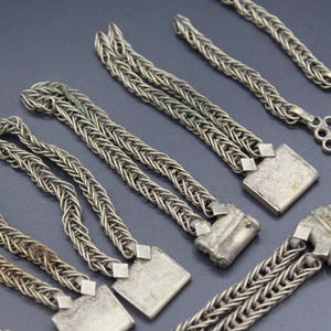 Pode incluir: Close-up de v&aacute;rias pe&ccedil;as de corrente de metal prateado. As correntes s&atilde;o intrincadamente tecidas, com elementos decorativos quadrados e circulares. As pe&ccedil;as est&atilde;o dispostas sobre uma superf&iacute;cie escura, mostrando seu artesanato detalhado.