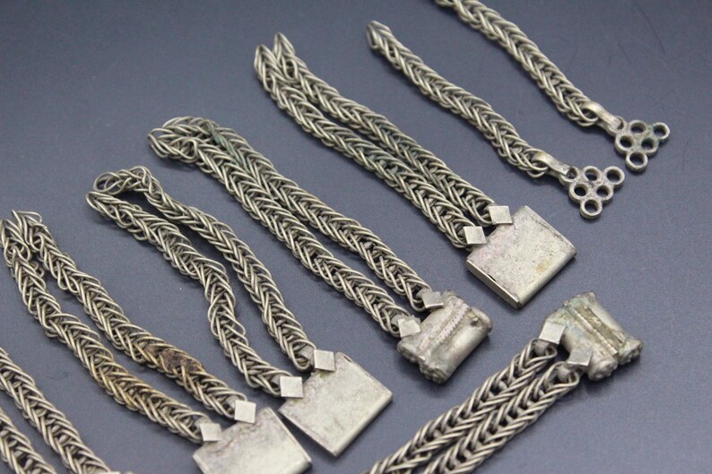 Pode incluir: Close-up de v&aacute;rias pe&ccedil;as de corrente de metal prateado. As correntes s&atilde;o intrincadamente tecidas, com elementos decorativos quadrados e circulares. As pe&ccedil;as est&atilde;o dispostas sobre uma superf&iacute;cie escura, mostrando seu artesanato detalhado.