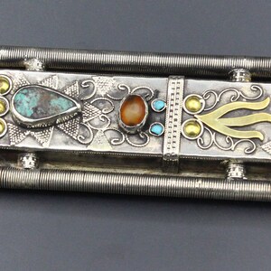 Puede incluir: Pieza decorativa de metal plateado ornamentada con piedras de color turquesa y &aacute;mbar. El dise&ntilde;o incluye detalles dorados y una forma central estilizada. Dos barras estriadas paralelas en la parte superior e inferior.