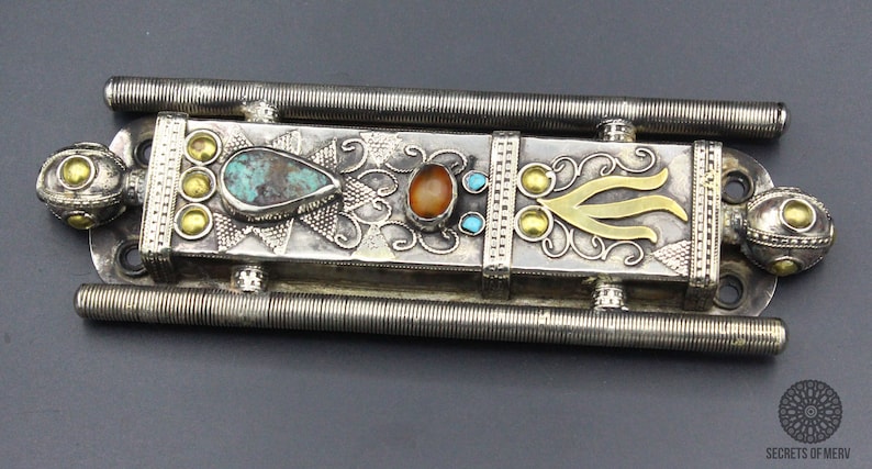 Puede incluir: Pieza decorativa de metal plateado ornamentada con piedras de color turquesa y &aacute;mbar. El dise&ntilde;o incluye detalles dorados y una forma central estilizada. Dos barras estriadas paralelas en la parte superior e inferior.